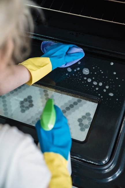Close-up of a person wearing yellow and blue rubber cleaning gloves, cleaning the glass surface of an oven door with a blue cloth. The oven, made of black material with a transparent glass panel, is s