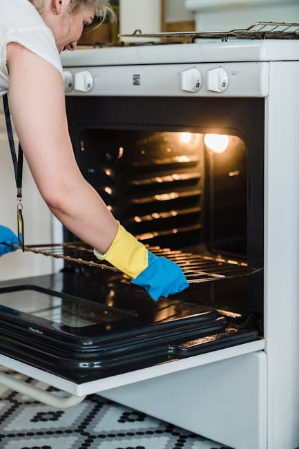 A woman with light-colored hair tied back, wearing a white t-shirt, a black apron, and yellow and blue rubber cleaning gloves, is performing surface cleaning on a white gas stove in a kitchen. She is 